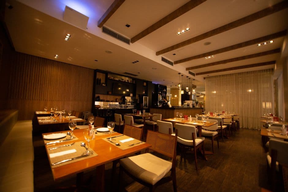 Warm, modern restaurant dining room with natural wood tables, soft overhead lighting, and a few guests sharing a meal in relaxed conversation — feels inviting and unhurried